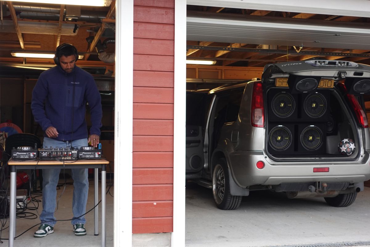 En mann står ved et DJ-miskebord. Det står en bil med store høytallere i bagasjen til høyre for ham.