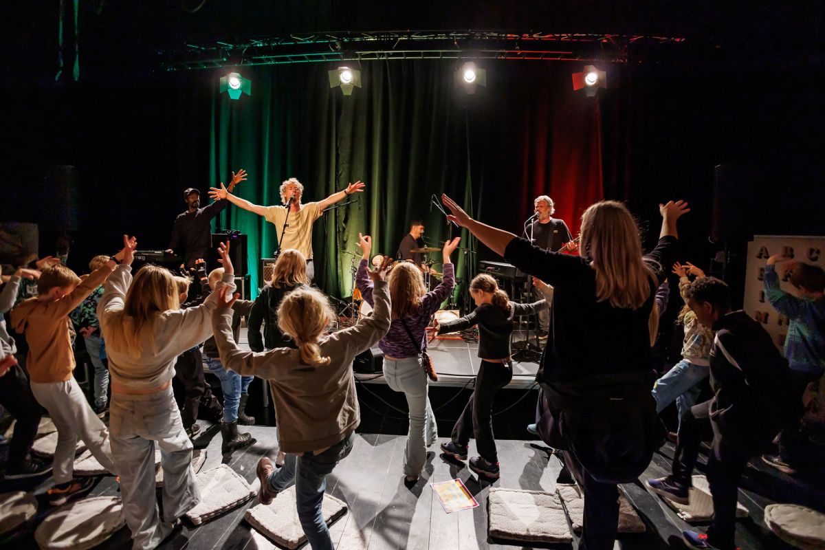 Konsertopptreden med energisk publikum. Forsanger på scenen med armene hevet. Fargerik belysning lyser opp band og publikum.