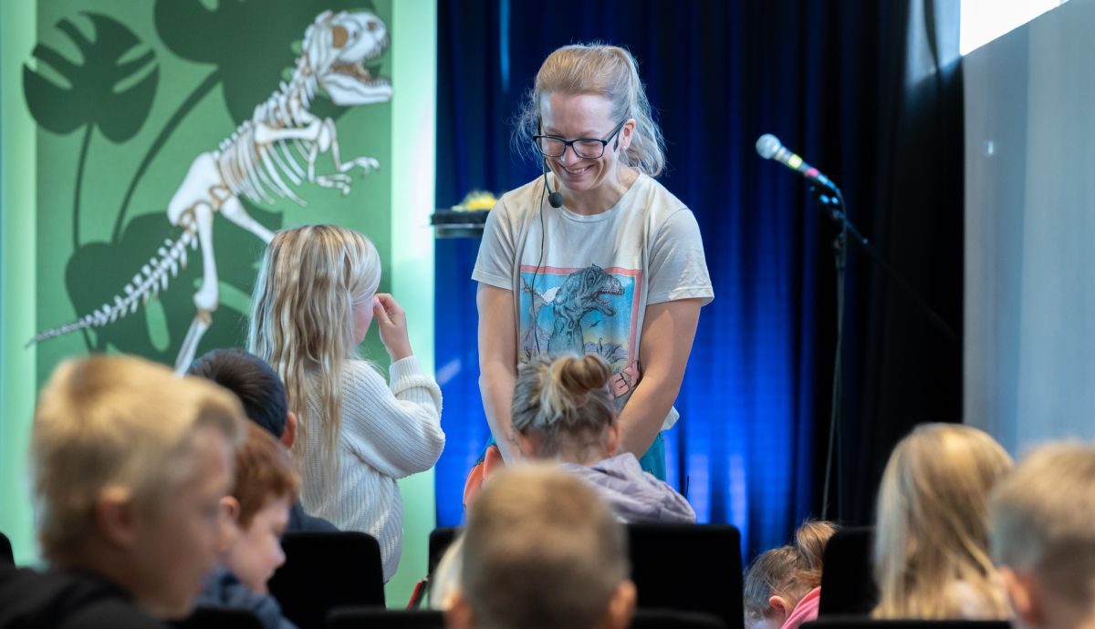 En kvinne med briller står på en scene foran flere barn og snakker med en jente.