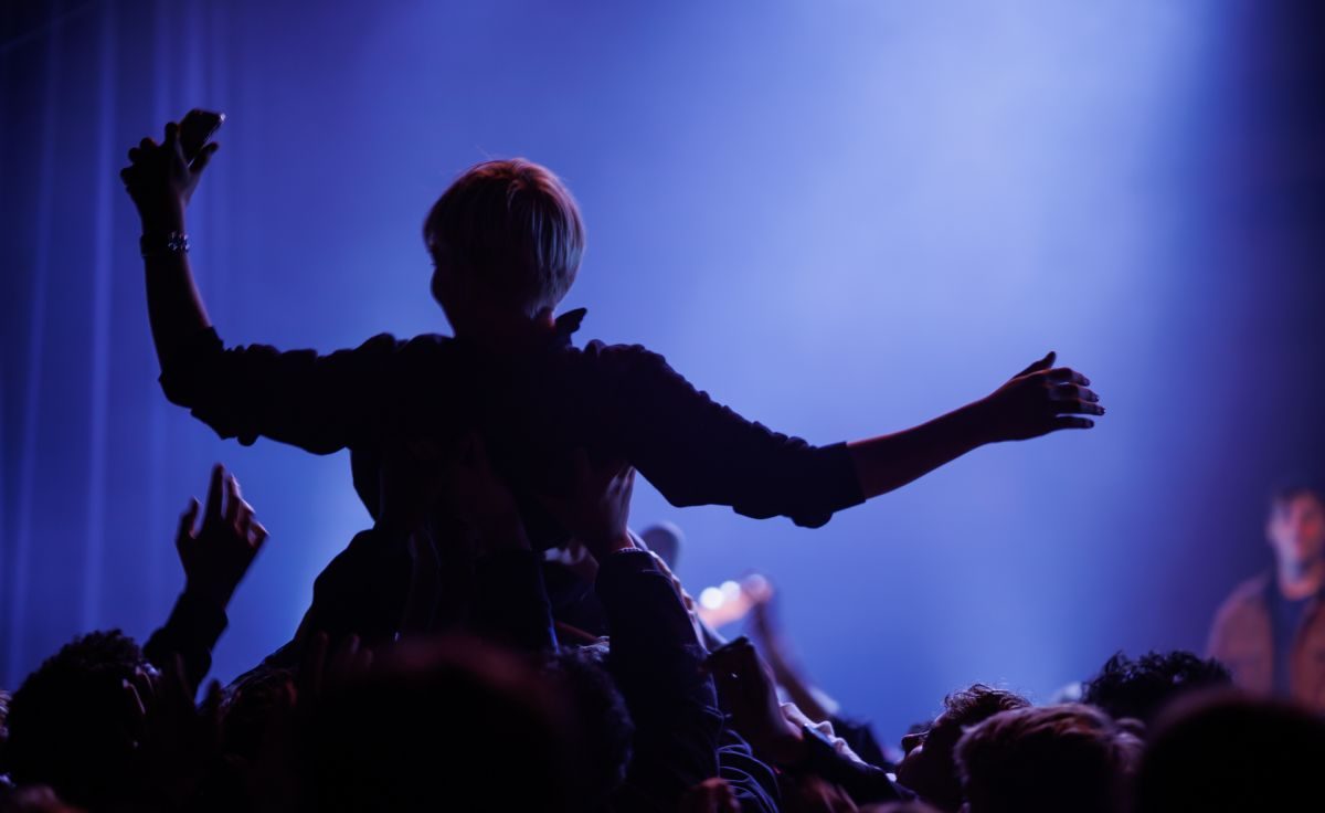 Silhuett av ungdom med utstrakte armer på toppen av publikum på konsert, badet i lilla-blå scenebelysning.
