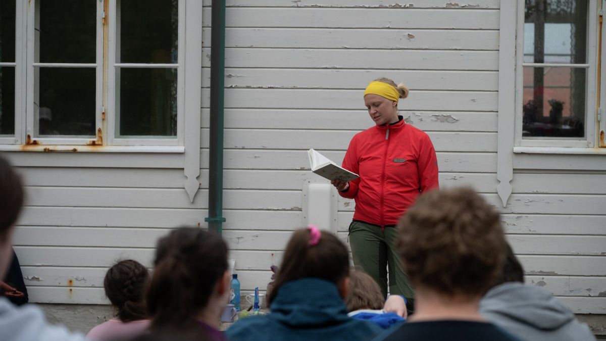 En kvinne i rød anorakk leser fra en bok til elever. De sitter ute.
