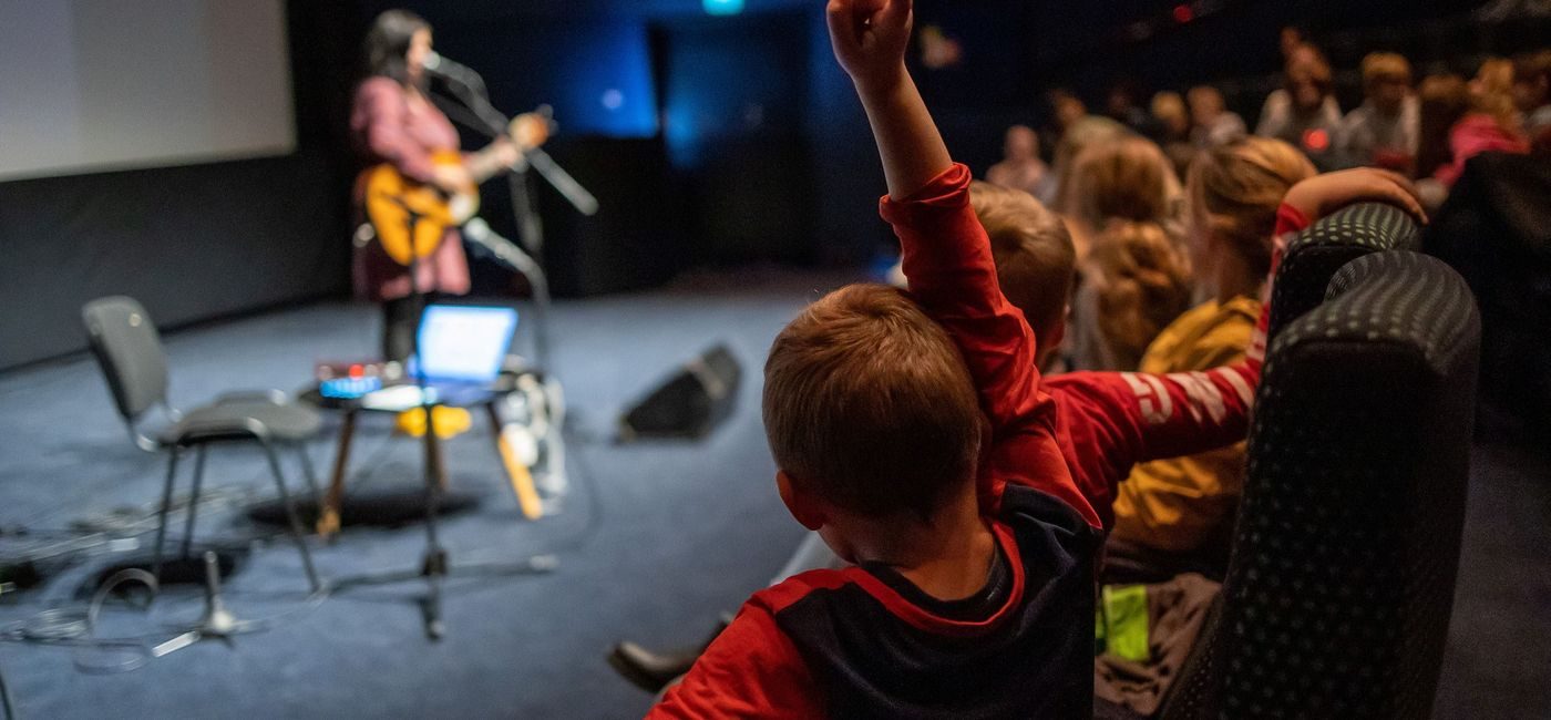 Et barn rekker opp hånden i publikum, på scenen står en dame som synger og spiller gitar