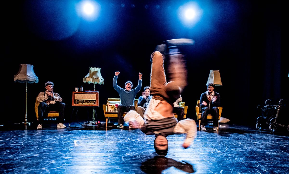 En mann danser breakdance. Hodet er i gulvet mens han snurrer. Bak ham sitter det fire menn i kostyme og heier. Bildet er tatt på forestilling fra en scene.