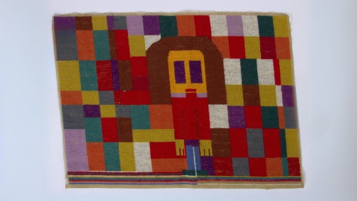 Verk av Wenche Nilsen. Broderi på stramei. Datering ukjent. Flerfarvet broderi med rektangulære og kvadratiske former i fargene blå, gul, oransj, lilla, blå, brun og hvit. I senter av broderi: En menneskeskikkelse bygget opp av rektangulære former. Med brunt hår, gult ansikt og gule hender, lilla øyne, rød overkropp, blå underkropp, grønne føtter. Langs underkant har kunstneren brodert inn vannrette tynne striper i fargene blå, lilla, oransj, rød, blå, grønn, gul, lys rosa, grå og rød til høyre i bildet. Til venstre i bildet er stripene hvit, gul, lys rosa, oransj, rød lilla, blå og grønn.