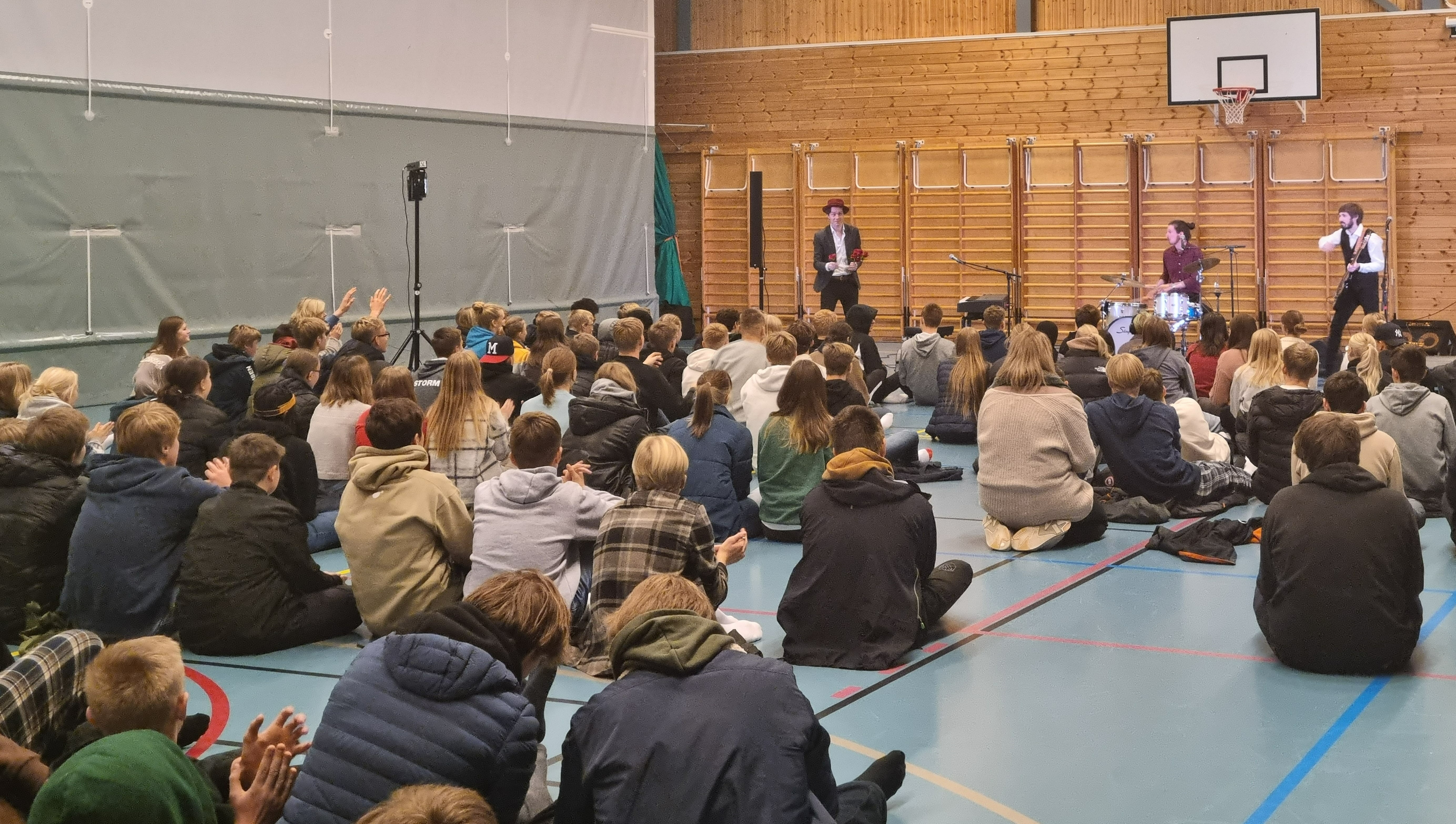 Konsert med Daniel Røssing Trio på en skole i Porsgrunn. Elever som sitter på gulvet i en gymsal, og tre musikere i enden som spiller trommer, bass og pianio.