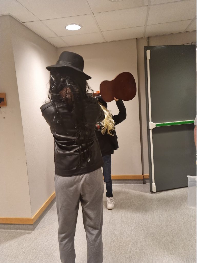 En elev med sort parykk og hatt tar bilde av en annen elev med lyst hår, hatt og gitar. Elev med sort parykk står med ryggen til kameraet og skjuler ansiktet til elev med lys parykk.