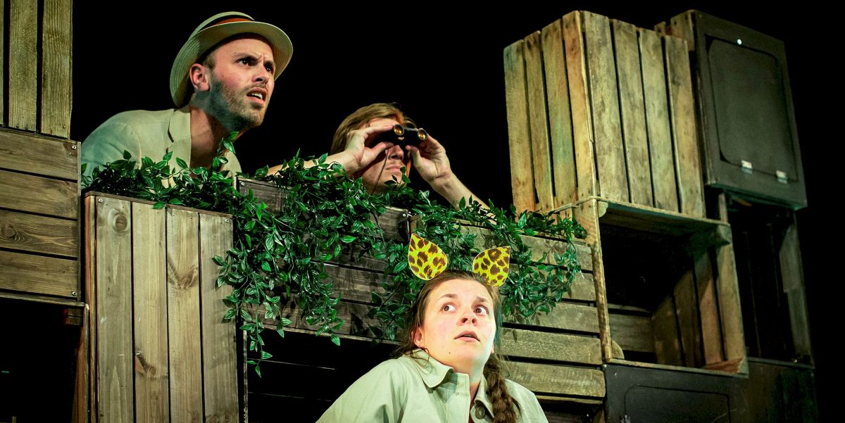 Scene frå scenekunstframsyning. Ein mann med hatt, ein med kikkert og ei kvinne med leopardøyrer på hovudet sit mellom tomme kassar og bladverk.